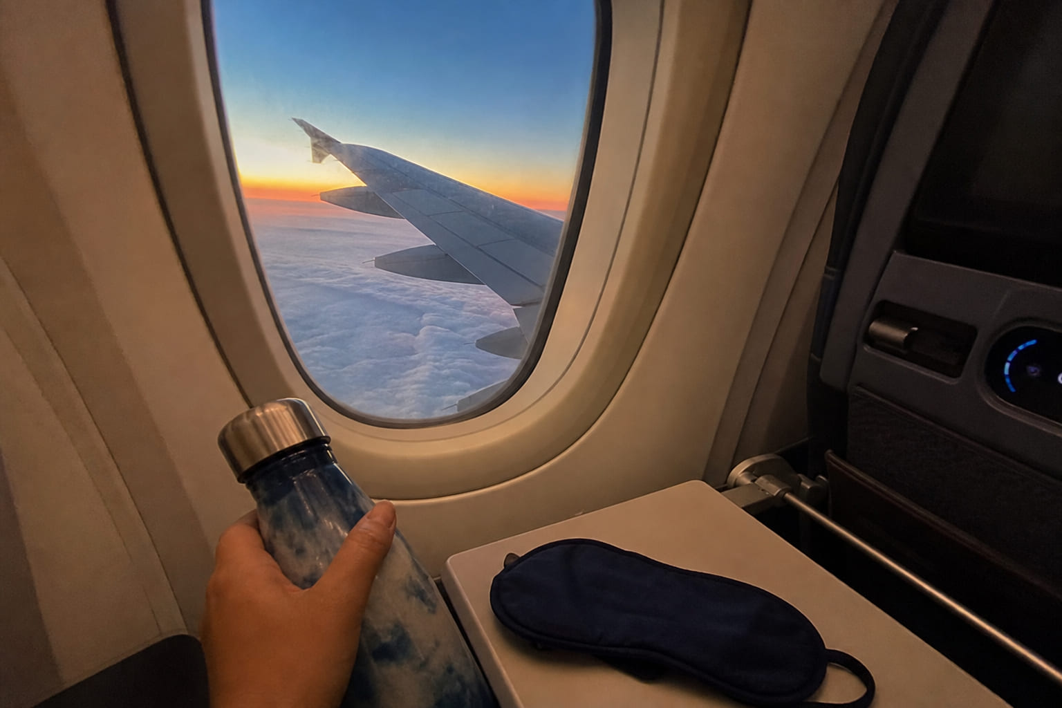 Phone snapshot from a window seat: airplane wing at dusk, sleep mask and water bottle on the tray table